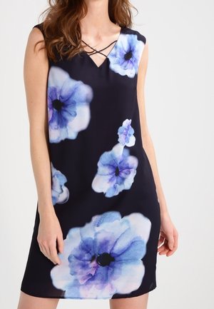 Sort ærmeløs kjole med store blå og lilla akvarelblomster og krydsdetalje ved V-halsen, båret af en kvinde.