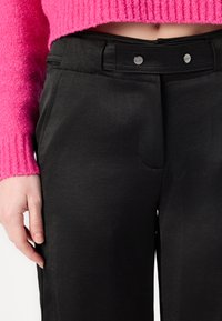 Μαύρο παντελόνι με λεία υφή, που διαθέτει δύο μπροστιν�ές τσέπες και ζώνη με μαύρο κουμπί ως διακόσμηση. Συνδυασμένο με ένα φωτεινό ροζ πουλόβερ.