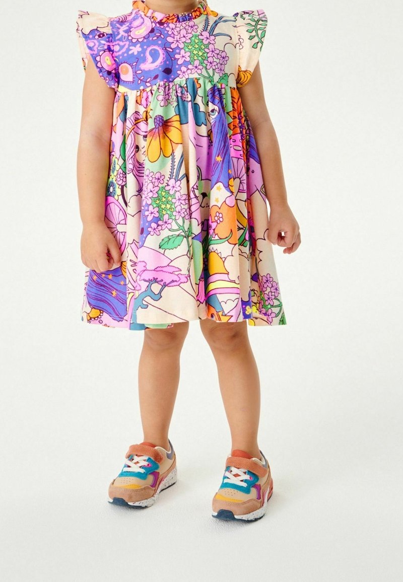 Enfant portant une robe colorée à imprimé floral et dessin animé avec des manches volantées et des baskets multicolores, debout sur fond blanc.