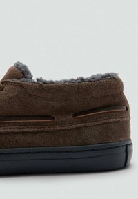 Hnědé semišové bačkory se šedou kožíškovou podšívkou, nízký profil, zaoblená špička a černá gumová podrážka. Obsahují dvojité dekorativní švy a výřezy.