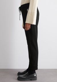 Črni tapered jogger hlače z raztegljivim pasom, stranskimi žeprsimi žepi in manšetami na gležnjih, v kombinaciji s črnimi loafersi in svetlo bež puloverjem.