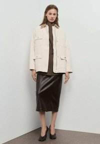Crème canvas jas met grote voorzakken, gelaagd over een bruine coltrui, gecombineerd met een donkerbruine leren pencil skirt en zwarte hakken.