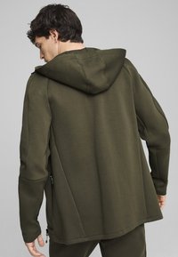 Donkergroene hoodie gemaakt van zacht materiaal, met een relaxte pasvorm, ritsen aan de zijkanten en textuuraccenten op de schouders.