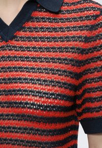 Haut à manches courtes en maille ouverte rayé rouge et noir avec col en V et peau visible en dessous.