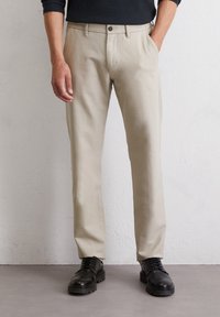 Beige trousers made of lightweight fabric featuring straight legs, belt loops, and a single button closure, paired with black lace-up shoes.