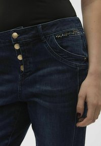 Los jeans de mezclilla azul oscuro presentan una cintura alta con cinco botones dorados y un bolsillo frontal adornado con lentejuelas doradas.