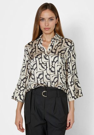 Frau trägt beige Seidenbluse mit schwarzem abstraktem geometrischem Muster und schwarze hoch taillierte Hose mit Gürtelring, steht vor weißem Hintergrund.