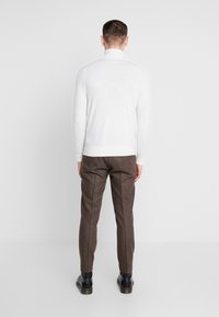 Man in een witte coltrui, bruine op maat gemaakte broek en zwarte schoenen, staat met zijn gezicht naar een effen witte achtergrond.