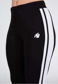 Leggings neri con strisce bianche laterali, realizzati in un materiale elastico, con un piccolo logo di gorilla bianco sulla coscia.