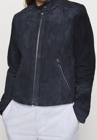 Tmavě modrá semišová bunda s vysokým límcem, předním zipem a dvěma bočními kapsami na zip. Hladký povrch a přiléhavý střih.