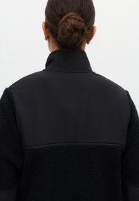 Czarna kurtka polarowa z gładkim górnym panelem. Część tułowia wykonana z teksturowanej, puszystej tkaniny. Wysoki kołnierz z zamkiem błyskawicznym i kontrastującymi fakturami.