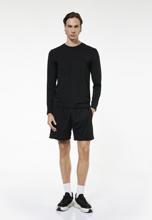 Młody mężczyzna stojący, ubrany w czarną koszulkę z długim rękawem, czarne szorty, białe skarpety oraz czarno-białe sneakersy na białym tle.