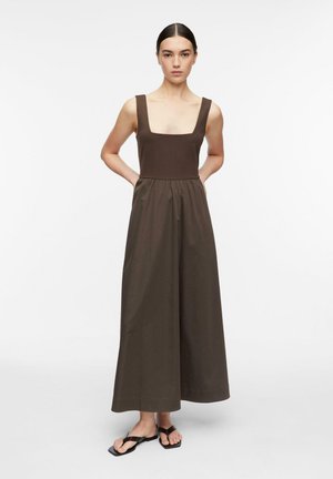 Kobieta o ciemnych włosach stoi z rękami za plecami, ubrana w brązowy, bezrękawnikowy kombinezon z szerokimi nogawkami i czarne sandały z kwadratowym noskiem.