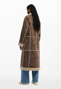 Cappotto lungo marrone con una finitura testurizzata, dettagli beige, e un rivestimento morbido. Presenta una forma squadrata e cuciture a griglia.