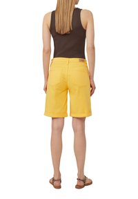 Shorts en denim jaune avec des poignets roulés, cinq poches et une étiquette en cuir au dos. Associés à un débardeur noir et des sandales marron.