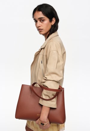 Mujer con cabello oscuro que lleva una chaqueta beige sostiene un gran bolso tote de cuero marrón sobre el brazo contra un fondo blanco.