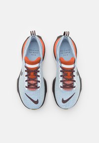 Světle modré sportovní boty mají texturovaný svršek, oranžové akcenty, černé podrážky a bílé tkaničky s logem Nike na boku.