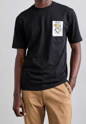 Homme portant un T-shirt noir à manches courtes avec un patch ourson Moschino sur la poche poitrine, et un pantalon beige avec le détail COUTURE! sur la poche.