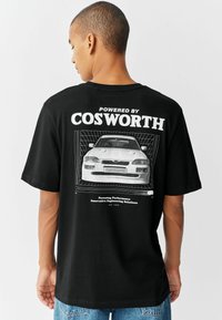 Camiseta de algodón negra con un gran estampado gráfico de un coche y el texto "Impulsado por Cosworth" en la parte trasera. Manga corta; corte relajado.