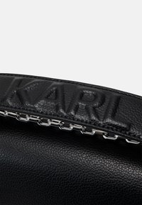 Juodas odinis rankinukas su iškilniu "KARL" logotipu, sidabrinės grandinėlės detalėmis, tekstūruota paviršiumi, fiksuota forma ir plonu dizainu.