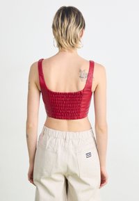 Top cropped em pele sintética vermelha com alças largas, apresentando uma textura franzida nas costas e um decote quadrado. Usado com calças de cintura alta em bege.