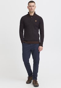 Brązowy sweter dzianinowy z kołnierzem na zamek, z prążkowaną teksturą i akcentem logo na piersi. Połączony z granatowymi spodniami i brązowymi butami.
