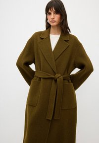 Olivgrön ullblandad kappa med sjalkrage, bälte vid midjan och två framfickor. Mjuk textur och avslappnad passform med långa ärmar.