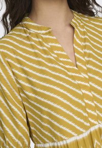 Gelbes Kleidungsstück mit weißen Zickzackstreifen, versehen mit einem halben Knopfverschluss am Ausschnitt und gerafften Ärmeln, hergestellt aus einem weichen, leichten Stoff.