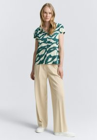 Blouse à motifs verts et crème avec des manches courtes, associée à un pantalon large beige et des baskets blanches. Tissu doux, coupe décontractée.