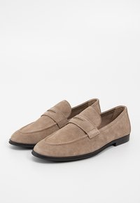 Paire de mocassins en daim marron clair avec semelles noires, design sans lacets, et couture décorative sur la surface supérieure.