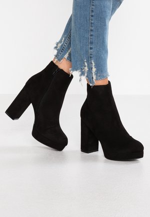 Jambes en jean bleu effiloché portant des bottines à plateforme en daim noir avec talons blocs et fermetures éclair latérales sur un sol blanc.