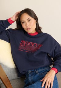 Marineblaues Sweatshirt mit roten Grafiken, auf denen "ATHLETICS CLUB" steht. Es hat einen gerippten Ausschnitt und gerippte Bündchen. Das Model trägt darunter ein rotes, rüschendes Hemd.
