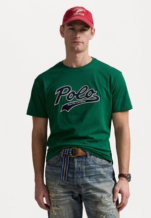 Camiseta de algodón verde con el logo de "Polo Ralph Lauren", combinada con vaqueros azul desgastados y un cinturón marrón con un acento a rayas. Gorra roja en la cabeza.