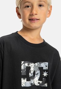 Camiseta de algodón negra con un gran logo de DC en gris con un patrón de salpicaduras. Cuello redondo y mangas cortas. Adecuada para uso casual.