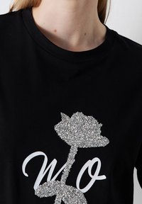Černé bavlněné tričko s texturovaným stříbrným třpytivým grafickým motivem růže a bílým nápisem "WO." Krátké rukávy a design s kulatým výstřihem.