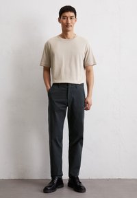 T-shirt en coton beige à manches courtes, associé à un pantalon vert foncé. Le modèle porte des bottes à lacets noires, les mains dans les poches, sur un fond neutre.