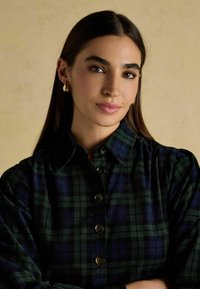 Zeleno-námornícku károvanú košeľu s golierom, zapínaním na gombíky a naberacím rukávom. Zlátnuté kruhové náušnice. Hladká textúra. Jemný pruhovaný vzor.