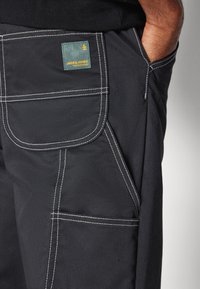 Czarne spodnie z białymi przeszyciami, z dużą kieszenią po bokach i zieloną metką z żółtym napisem z tyłu. Wytrzymały materiał.