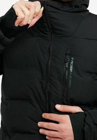 Veste noire rembourrée avec col haut et fermeture éclair sur le devant, arborant un logo sur la poitrine et des ouvertures pour les pouces aux poignets pour plus de chaleur.
