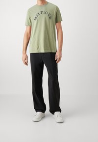 Grøn bomulds t-shirt med "HILFIGER" printet med mørke bogstaver, parret med sorte bukser og hvide sneakers. Casual pasform, rund halsudskæring.