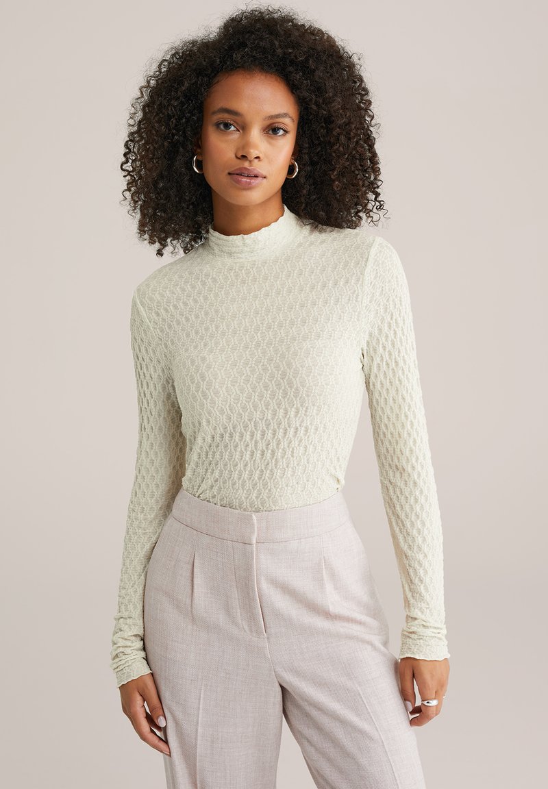 Jasnozielony top z długim rękawem wykonany z teksturowanej tkaniny z wysokim dekoltem, zestawiony z jasno-różowymi, dopasowanymi spodniami.