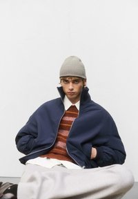 Tengerész bomber dzseki magas gallérral, barna csíkos kötött pulóver, fehér ing, szürke nadrág és bézs kötött sapka. Simaszerű textúra és laza illeszkedés.