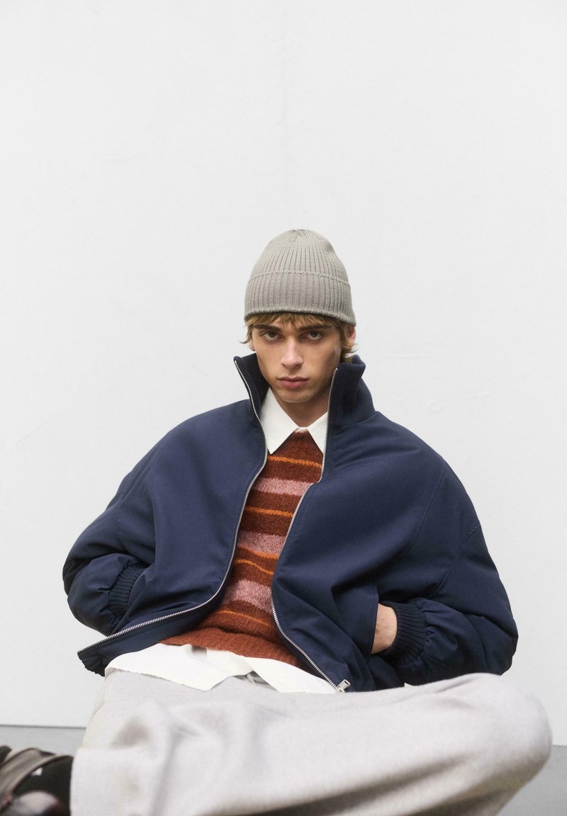 Tengerész bomber dzseki magas gallérral, barna csíkos kötött pulóver, fehér ing, szürke nadrág és bézs kötött sapka. Simaszerű textúra és laza illeszkedés.