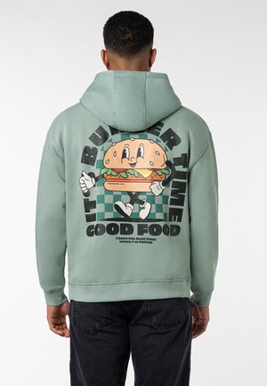 Grüne Kapuzenjacke aus Baumwollmischung mit einem großen Cartoon-Burger-Design auf der Rückseite, ergänzt durch fett gedruckten Text und Schachbrettakzente.