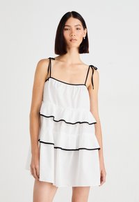 Robe blanche avec des bretelles réglables, comportant des accents de volants noirs au bas et à la poitrine. Confectionnée en tissu léger avec un design à volants.