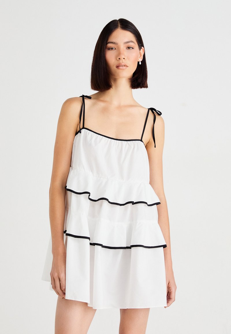 Robe blanche avec des bretelles réglables, comportant des accents de volants noirs au bas et à la poitrine. Confectionnée en tissu léger avec un design à volants.
