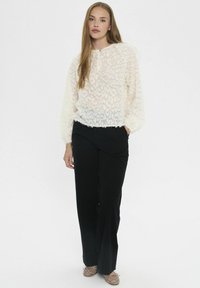 Donna con i capelli lunghi che indossa una blusa bianca a trama, pantaloni neri a gamba larga e ballerine tan con borchie, in piedi su uno sfondo semplice.