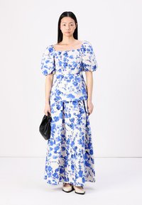 Robe florale avec des motifs bleus et blancs, manches bouffantes, encolure carrée et jupe longue fluide. Sac noir et chaussures bicolores.