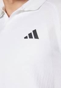 Camisa polo blanca con un acabado texturizado. Presenta un logo negro con tres rayas ascendentes en la parte izquierda del pecho. Diseño de collar clásico.