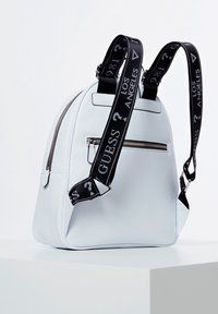 Bílý kožený batoh s zaobleným designem, vybavený nastavitelnými černými popruhy s logem, přední kapsou na zip a texturovaným povrchem.